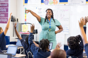 picture of female teacher smiling and pointing at students as students raise their hands
